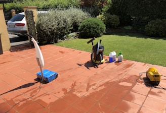 A sunlit patio with terracotta tiles is adorned with cleaning equipment, including a wet and dry vacuum, containers of cleaning solutions, and a pressure washer. A portion of a white car is visible near a hedge-lined driveway, while lush green vegetation and a manicured lawn provide a backdrop.