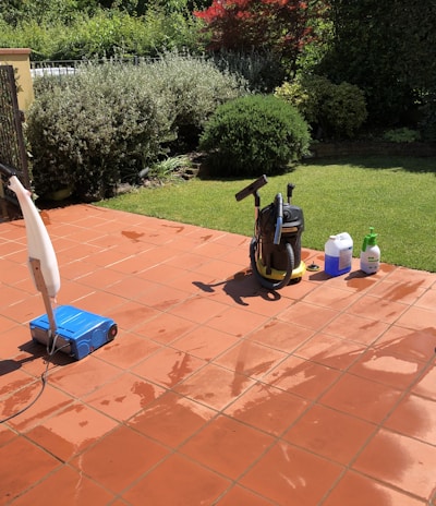 A sunlit patio with terracotta tiles is adorned with cleaning equipment, including a wet and dry vacuum, containers of cleaning solutions, and a pressure washer. A portion of a white car is visible near a hedge-lined driveway, while lush green vegetation and a manicured lawn provide a backdrop.