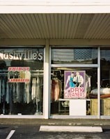 A storefront for a dry cleaning business with large glass windows displaying various signs and clothing racks with garments hung inside. The signage includes 'Nashville's Shirts Laundered' and services like spotting, drapery, silk and wool care. The central window features a colorful poster of a man and woman labeled 'Dry Cleaners.' An American flag is visible in the reflection on the glass.