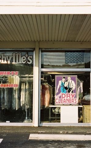 A storefront for a dry cleaning business with large glass windows displaying various signs and clothing racks with garments hung inside. The signage includes 'Nashville's Shirts Laundered' and services like spotting, drapery, silk and wool care. The central window features a colorful poster of a man and woman labeled 'Dry Cleaners.' An American flag is visible in the reflection on the glass.