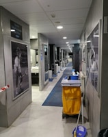 A hallway with gray tiles and blue carpet features cleaning equipment, including a yellow cleaning cart and a mop. There are black and white sports photographs on the walls and a door leading to a restroom area. Overhead, there are ceiling lights providing illumination.