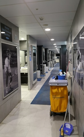 A hallway with gray tiles and blue carpet features cleaning equipment, including a yellow cleaning cart and a mop. There are black and white sports photographs on the walls and a door leading to a restroom area. Overhead, there are ceiling lights providing illumination.