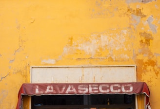 A vibrant yellow, weathered wall with a green, peeling wooden shutter above a storefront. The faded maroon awning displays the word 'LAVASECCO'. Below, the entrance reveals a glimpse into a shop with cleaning equipment visible and informational posters displayed on the glass.