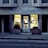 A dry cleaning shop named 'HLP Cleaners' is located on a dimly lit street. The store has a prominent sign above the entrance, and its windows are illuminated with a warm yellow light. A sign on the door lists services such as dry cleaning, laundry, and suede & leather cleaning. The building exterior is simple with a combination of dark tiles and light-colored upper walls. The entrance is flanked by two closed doors, likely of neighboring establishments.
