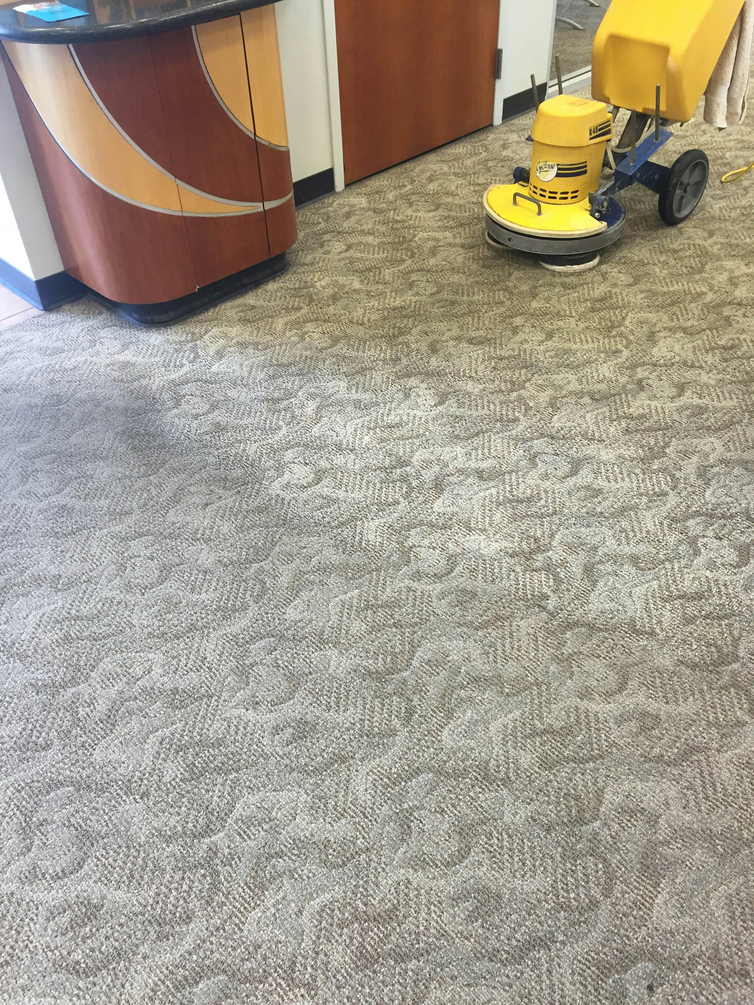 A patterned carpet covers most of the floor, with a yellow floor cleaning machine partially visible on the right. The pattern on the carpet appears to be a mix of geometric and abstract shapes. A section of a wooden counter with a curved design is on the left side.